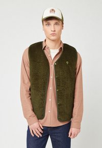 Gilet di velluto a coste verde oliva con righe verticali, bottoni frontali e un piccolo logo, indossato sopra una camicia a quadri rosa e jeans scuri.