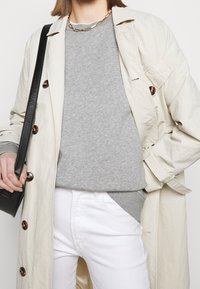 Gabardina beige claro con botones marrones brillantes, sobre un suéter gris. Jeans blancos y un bolso negro con correa de hombro.