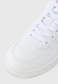 Λευκό δερμάτινο αθλητικό παπούτσι με λεία υφή, στρογγυλεμένη μύτη, οπές στην άκρη της μύτης και επίπεδα λευκά κορδόνια.