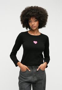 Čierny dlhý rukáv s rebrovým vzorom a malou ružovou srdcovou grafikou na hrudi. V kombinácii so šedými džínsami s jemnými vzormi.