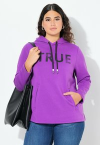 Sudadera morada con estampado negro de "TRUE", capucha con cordón, bolsillos tipo canguro y tejido texturizado. Combinada con jeans azules y un bolso negro.