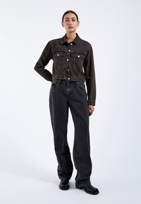 Giacca di denim marrone con due tasche frontali, chiusura con bottoni e design cropped, abbinata a jeans scuri a gamba larga e stivali neri.