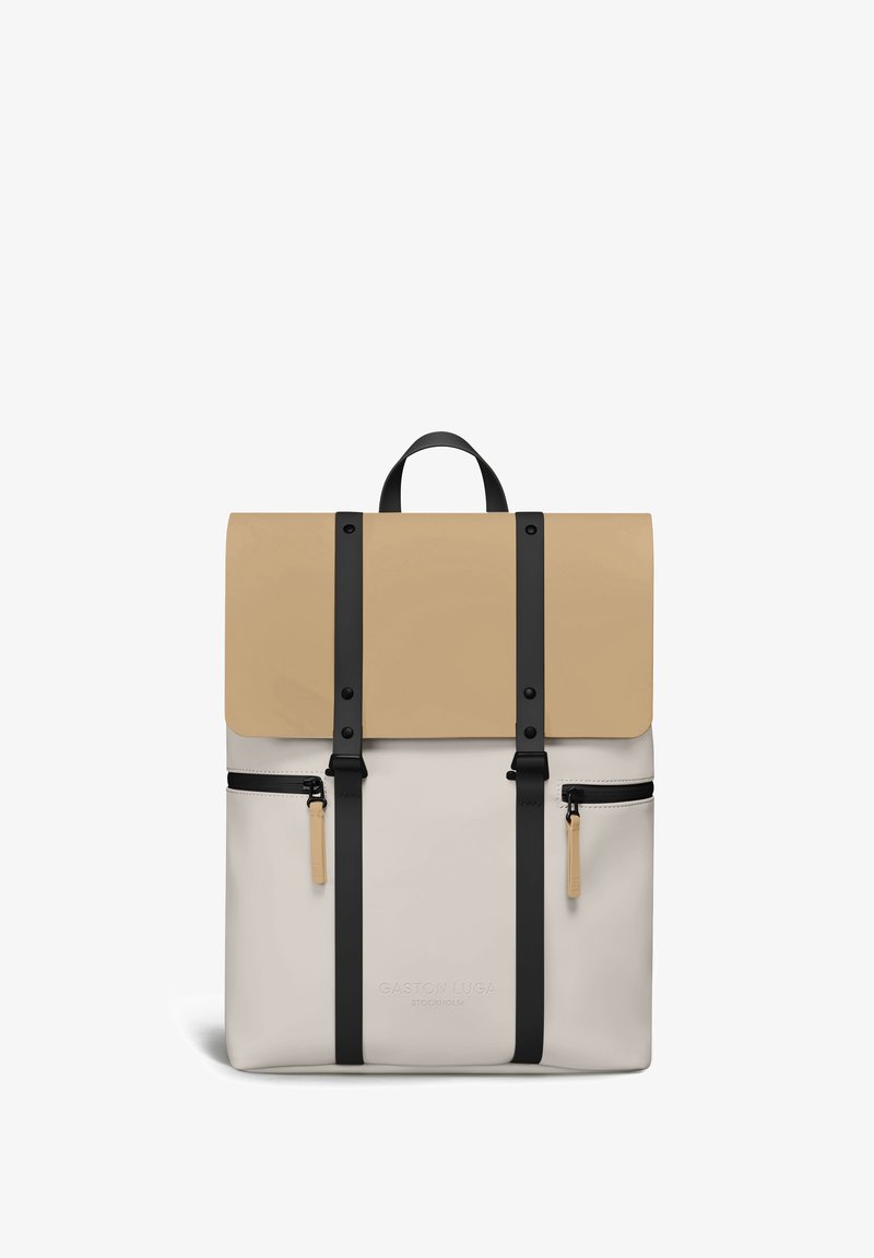 Sac à dos beige et crème avec des sangles verticales noires, deux poches avant zippées et une poignée supérieure, signé Gaston Luga Stockholm.