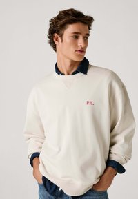 Felpa beige chiaro con scollo tondo, caratterizzata da un piccolo logo rosso "PJL". Indossata sopra una camicia di jeans scura con colletto. Vestibilità casual.