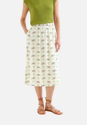Mujer con falda midi blanca con estampado de elefante y palmera, top verde sin mangas y sandalias envueltas de color beige, de pie con las manos en los bolsillos.