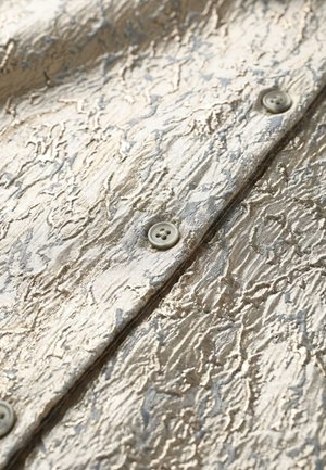 Close-up van glanzende, gestructureerde beige stof met grijze abstracte patronen en viergaatjes knopen genaaid langs een centrale naad.