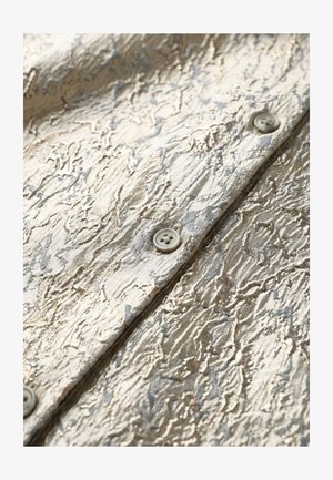 Close-up van glanzende, gestructureerde beige stof met grijze abstracte patronen en viergaatjes knopen genaaid langs een centrale naad.