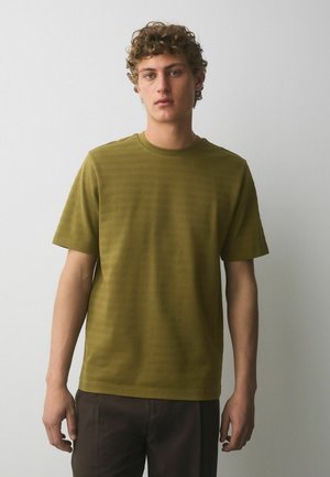 Młody mężczyzna z kręconymi włosami, ubrany w oliwkowo zieloną, teksturowaną koszulę z krótkim rękawem i ciemnobrązowe spodnie, stojący na tle gładkiej białej ściany.