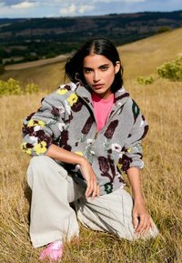 Fleece zip-up jacket with floral pattern in gray, yellow, and maroon; paired with light beige pants and pink shoes. Natural outdoor setting.