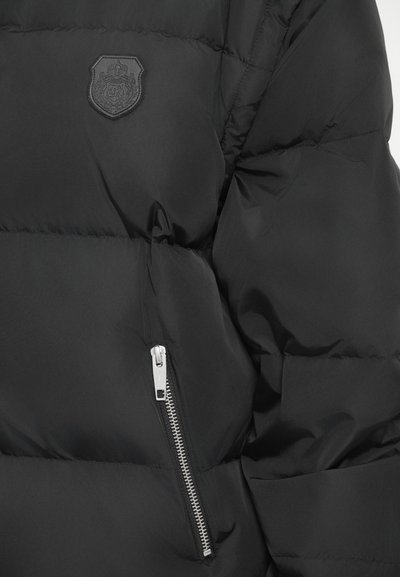 Μαύρο μπουφάν πάνας με λογότυπο σχήματος ασπίδας, κατακόρυχη τσέπη με φερμουάρ και ασημένια μεταλλικά στοιχεία, καθώς και επένδυση με καπιτονέ υφή για μόνωση.