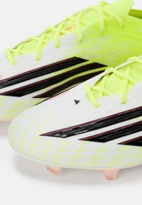 Λευκά ποδοσφαιρικά παπούτσια με μαύρες και κόκκινες ρίγες, διαθέτουν υφή πάνω μέρος και κορδόνια νέον κίτρινου χρώματος. Διάφανοι πλαστικοί καρφίτσες στη σόλα.