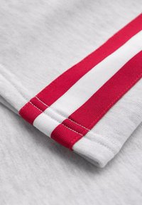 Tela de camisa gris con una textura suave, que presenta un detalle de rayas rojas y blancas a lo largo del dobladillo de la manga. Costura limpia visible.