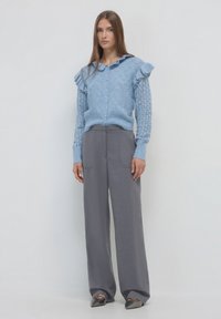 Donna in piedi che indossa un cardigan di maglia blu con spalle arricciate, pantaloni grigi a gamba larga e scarpe nere a punta su uno sfondo neutro.