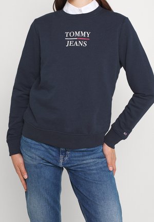 Persoon draagt een marineblauwe sweatshirts met witte "TOMMY JEANS" tekst en onderstreping, gecombineerd met een blauwe spijkerbroek en een witte overhemd met kraag eronder.
