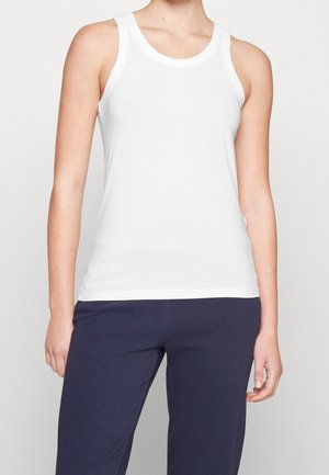 Personne portant un débardeur blanc uni sans manches et un pantalon bleu foncé, debout devant un fond gris clair.