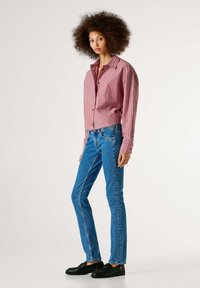 Rød- og hvitstripet skjorte med knapper og lange ermer, kombinert med blå, tettsittende jeans og svarte loafers. Glatte teksturer og avslappet design.