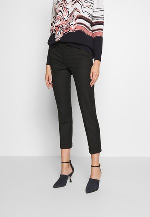 Vrouw draagt zwarte cropped broek, zwarte hakken met enkelbandje en een meerkleurige trui met lange mouwen en een abstract patroon.