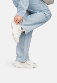 Baskets blanches épaisses avec des détails en mesh texturé et en cuir, ornées d'accents dorés, associées à un pantalon en denim bleu clair.