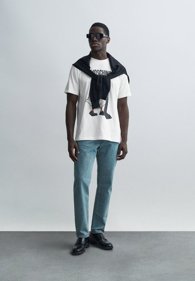 Valge t-särk graafilise disainiga, hele sinised teksad, mustad kingad ja must kampsun, mis on õlgadele visatud. Päikeseprillid täiendavad välimust.