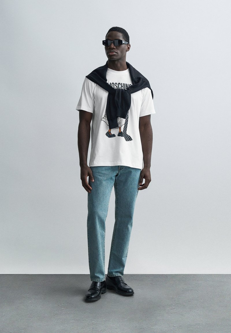 Weißes T-Shirt mit Grafikdesign, hellblaue Jeans, schwarze Schuhe und ein schwarzer Pullover, der über die Schultern gelegt ist. Sonnenbrille rundet das Outfit ab.