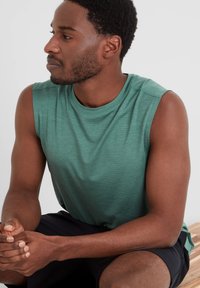 Sleeveless green athletic shirt made of textured fabric, featuring a round neckline, worn with dark shorts, seated on a wooden surface.