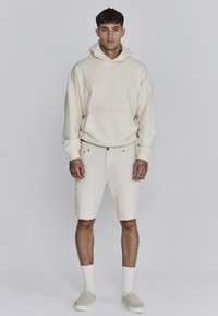 Beige hoodie met "SIKSILK" logo, gecombineerd met lichtbeige denim shorts met een ruwe zoom, voorzakken en casual instapschoenen.