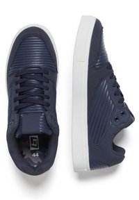 Navy sneakers feature a textured synthetic upper with ribbed accents, round laces, and a white rubber sole. Size 44 visible inside.
