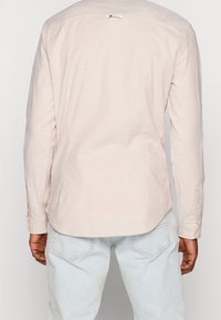 Man in een lichtbeige langslav shirt en lichtblauwe jeans, gezien vanaf de achterkant tegen een effen achtergrond.