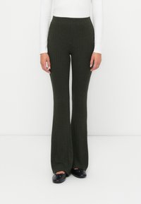 Geribbelde donkergroene uitlopende broek met een hoge taille. De stof heeft een gestructureerde afwerking. Draagt met een witte blouse met lange mouwen en zwarte flats.