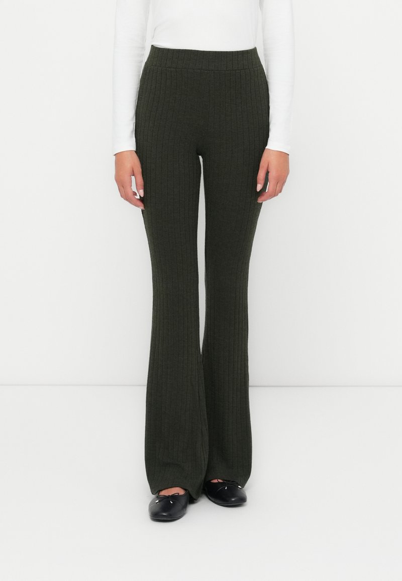 Geribbelde donkergroene uitlopende broek met een hoge taille. De stof heeft een gestructureerde afwerking. Draagt met een witte blouse met lange mouwen en zwarte flats.