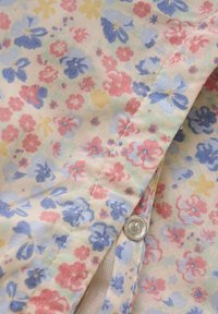 Stof met bloemenpatroon met kleine roze, blauwe en gele bloemen op een crèmekleurige achtergrond; bevat een zilveren drukknoop en zichtbare stiksels.
