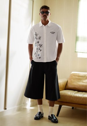 Uomo che indossa una camicia bianca a maniche corte con disegno floreale e la scritta "All Things Gone", pantaloncini neri larghi, scarpe nere, calzini bianchi e occhiali da sole al chiuso.