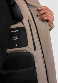 Chaqueta exterior beige con un forro acolchado negro, que cuenta con un bolsillo con cremallera y una etiqueta de marca que muestra "KHUJOTM". Textura de tela suave.
