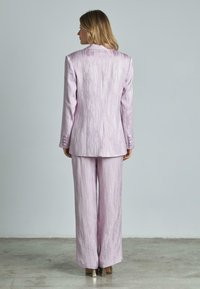 Completo viola con una finitura lucida e strutturata; giacca su misura e pantaloni a gamba larga. Bottoni chiusi sulla giacca.