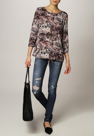 Mujer con blusa de manga larga floral, jeans ajustados rasgados, zapatos planos negros, sosteniendo un bolso grande tote negro, de pie frente a un fondo neutro.