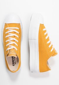 Keltaiset kangaskengät valkoisella kärkiosalla ja pohjalla, joissa on valkoiset nauhat ja teksturoitu sivu. Huomattava brändäys kielen kohdalla.