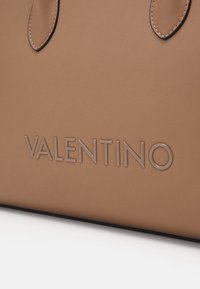 Brun läderhandväska med präglad "VALENTINO"-logotyp. Har en slät yta och kontrasterande sömmar längs handtagen.