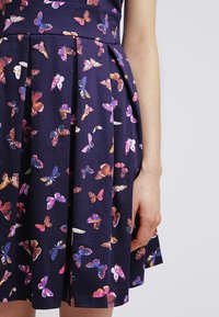 Marinblå kjol med veck som har ett färgglatt fjärilsmönster, visad på en persons nedre torso och högra arm mot en ljus bakgrund.