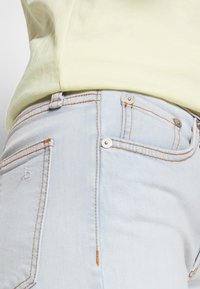 Jean en denim bleu clair avec coutures orange et rivets argentés, porté avec une chemise jaune pâle rentrée à la taille.