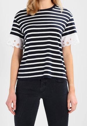 Frau trägt ein T-Shirt mit horizontalen dunkelblau-weißen Streifen und weißen, mit Muschelkante verzierten Ärmeln mit Stern-Ausschnitten, kombiniert mit schwarzen Hosen.