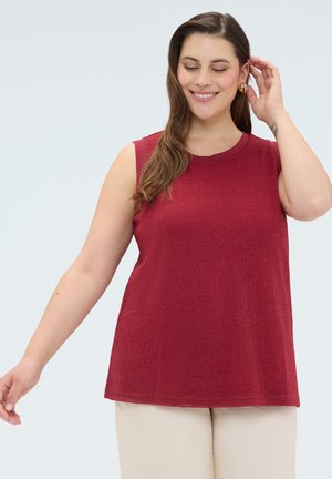 Kvinde med langt brunt hår iført ærmeløs rød top og beige bukser, smilende med lukkede øjne, hånd berører håret.