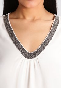 Blusa bianca con profondo scollo a V adornato con un bordo diperline grigie. Tessuto morbido con pieghe sul colletto che aggiungono texture e dettagli.