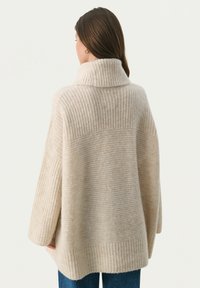 Beige oversized sweater med en ribbet høj krave og kontrasterende ribbede teksturer. Blødt strikket materiale med sidelufthuller og forlængede ærmer.