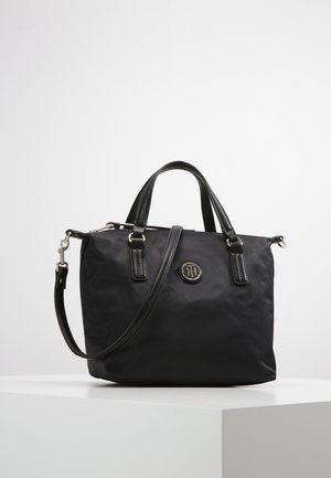 Black handbag with dual top handles, detachable shoulder strap, and round metallic logo emblem on front, displayed on a white pedestal.