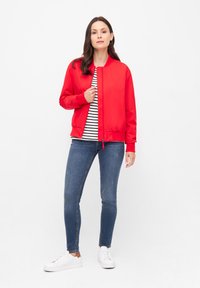 Röd bomberjacka med en slät, glänsande yta; ståkrage; framtida dragkedja; kombinerad med en randig skjorta, blå jeans och vita sneakers.