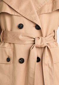 Beige trenchcoat met een brede kraag, dubbelzijdig ontwerp, zwarte knopen en een strikceintuur in de taille. Heeft twee voorkeurszakken.