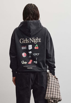 Persona che indossa una felpa nera con cappuccio con testo "Girls Night" e icone di rossetto, ciliegie, tacchi alti, occhiali da sole, e tiene una giacca a quadri.