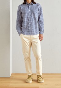 Blauw-witte geruite blouse met lange mouwen, beige broek en beige schoenen, staande op een houten vloer tegen een lichtgekleurde muur.