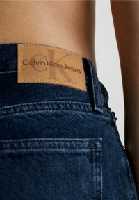 Närbild av en person som bär blå denimjeans från Calvin Klein med en beige läderlogg som är sydd på midjebandet.