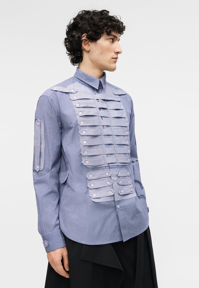 Homme faisant face à droite portant une chemise bleue à boutons avec des bretelles en tissu superposé et des boutons sur la poitrine et les manches, associée à un pantalon noir.